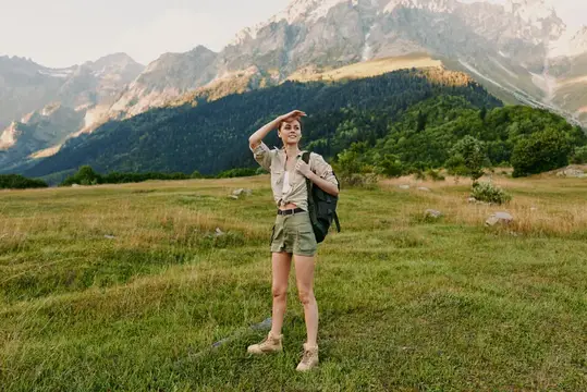 Женщина с рюкзаком на широком лугу на фоне возвышающихся вдалеке вершин, что наводит на мысли об исследованиях, свободе и связи с природой в условиях активного отдыха