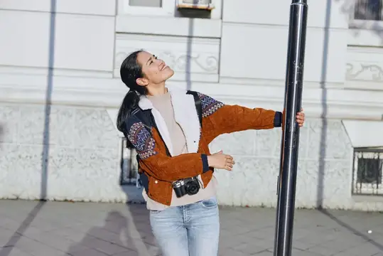 Молодая женщина с фотокамерой весело обнимает фонарный столб на улице