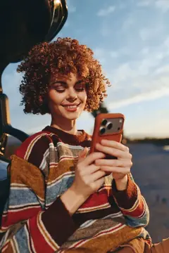 Рыжеволосая девушка с кудрями улыбается, глядя в смартфон на улице