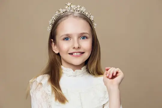 Улыбающаяся девочка в короне и белом платье на бежевом фоне