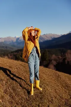 Женщина улыбается, показывая зубы, в полный рост поднимает руки вверх от счастья, идет по холму и смотрит на горы