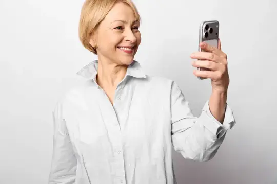 Улыбающаяся блондинка в белой рубашке делает селфи на смартфон