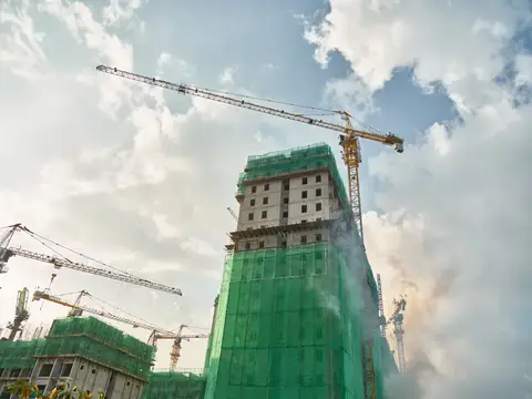 Современная строительная площадка с большими кранами и строительными лесами, покрытыми зелёной сеткой, на фоне пушистых облаков