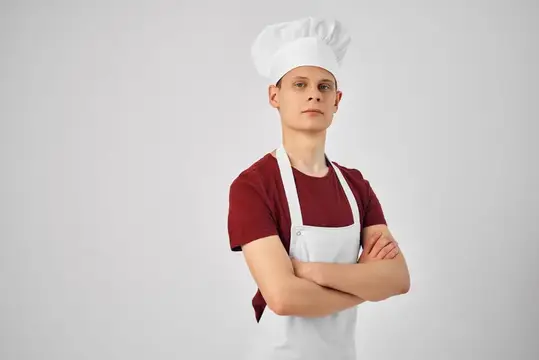 Шеф-повар в белом фартуке сложил руки перед собой, что подчеркивает его уверенность в себе и профессионализм