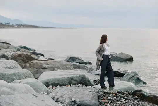 Женщина в плаще стоит на камнях у моря в пасмурный день