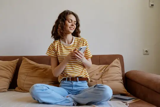 Улыбающаяся кудрявая девушка со смартфоном сидит на диване с подушками
