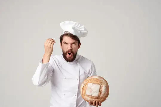 Бородатый пекарь в колпаке грозит кулаком, держа круглый хлеб