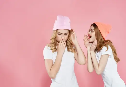 Две молодые женщины в шапках шепчутся на розовом фоне