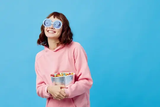 Улыбчивая девушка в розовом худи и круглых очках с миской конфет