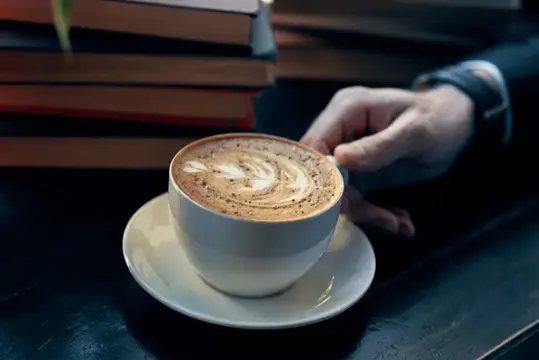 Мужская рука тянется к чашке кофе с пенкой рядом со стопкой книг