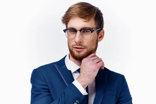 Молодой мужчина в очках и синем костюме задумчиво касается бороды
