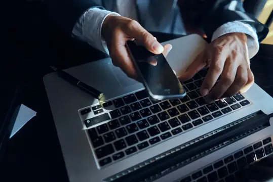 Мужчина в рубашке держит смартфон над клавиатурой ноутбука