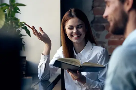 Улыбающаяся девушка с книгой беседует с мужчиной в офисе