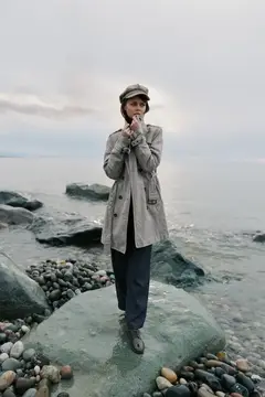 Девушка в плаще и кепке стоит на камне у моря
