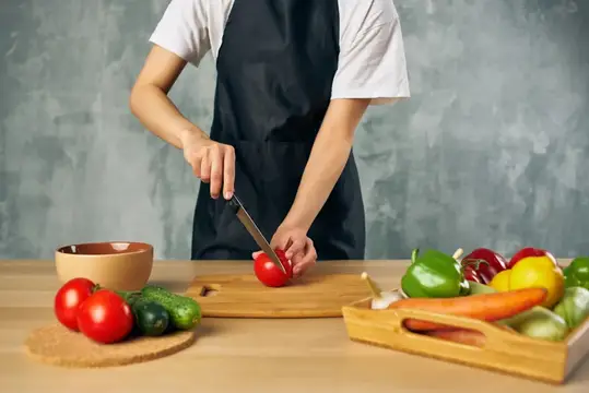 Повар в чёрном фартуке режет помидор на кухне
