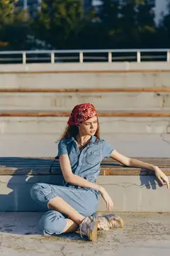 Женщина, одетая в современную джинсовую уличную моду бохо-вестерн, сидит расслабленно на бетонных ступеньках с красной банданой, в повседневной обуви; естественный солнечный свет подчёркивает её…