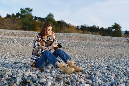 Фотограф на отдыхе на галечном пляже с ручной камерой, одетый в клетчатую рубашку и прочные ботинки, наслаждающийся природой и тихим берегом при ярком свете