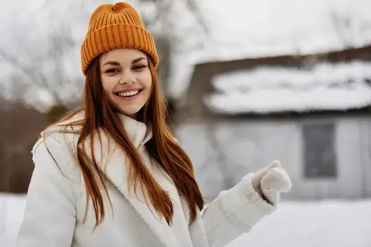 Улыбающаяся девушка в оранжевой шапке зимой на фоне дома