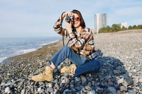 Улыбающаяся девушка в клетчатой рубашке фотографирует на каменистом берегу моря