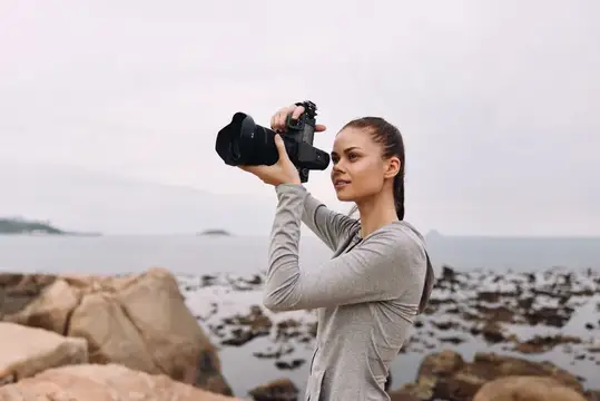 Целеустремленная молодая женщина в серой футболке с длинным рукавом фотографирует красивый прибрежный пейзаж, отражая свою любовь к фотографии в тихий день на пляже