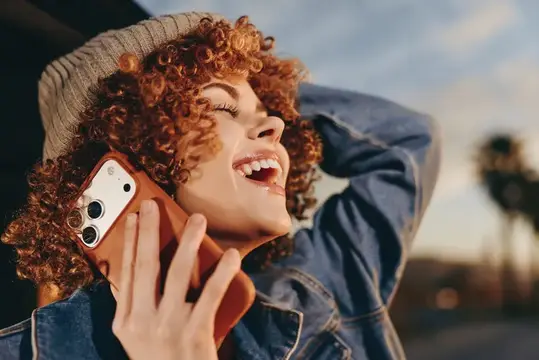 Женщина отдыхает на улице, улыбается и смеется, рыжие вьющиеся волосы под вязаной шапкой, джинсовая куртка, смартфон у уха, разговор по телефону, сияние заката, дружелюбное расслабленное настроение…