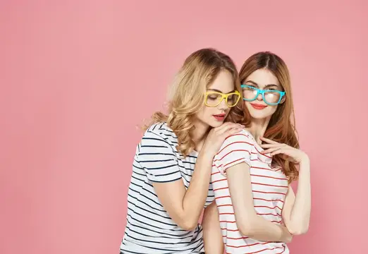 Две молодые женщины в ярких очках обнимаются на розовом фоне
