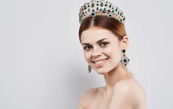 Улыбающаяся шатенка в роскошной короне с изумрудами на светлом фоне