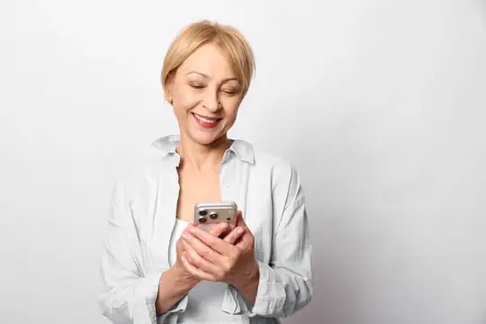Улыбающаяся блондинка в белой рубашке смотрит в смартфон
