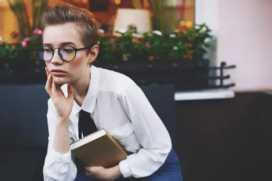 Образованная женщина с книгой и в очках сидит в кресле на веранде кафе