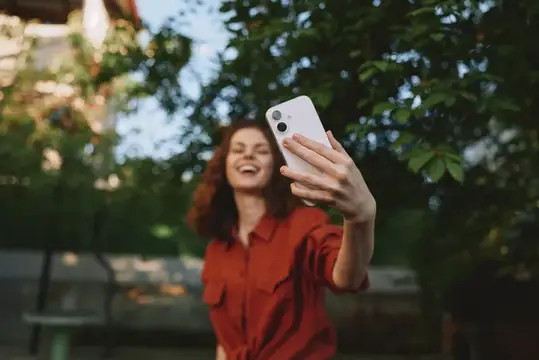 Рыжеволосая женщина в красной рубашке смеётся, делая селфи на смартфон в саду