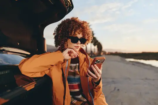 Рыжеволосая девушка в солнечных очках смотрит в телефон у машины