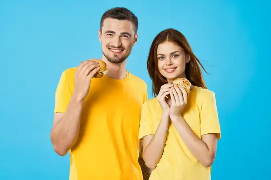 Улыбающиеся парень и девушка в жёлтых футболках едят булочки на синем фоне