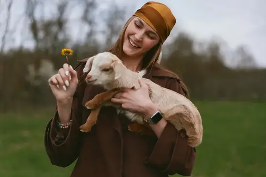Молодая женщина в коричневом пальто и оранжевом платке нежно держит козленка и протягивает ему желтый цветок, стоя на травянистом поле с размытыми деревьями на заднем плане