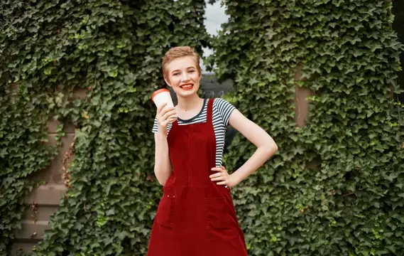 Симпатичная женщина с чашкой кофе рядом с зелёными листьями кустов на улице