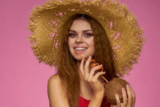 Улыбающаяся девушка в соломенной шляпе с кокосом и трубочкой на розовом фоне