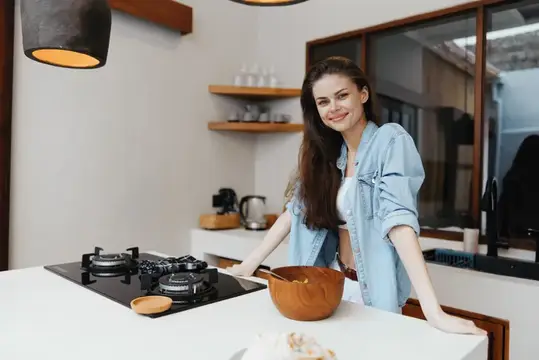 Веселая молодая женщина в светлой джинсовой рубашке стоит на современной кухне и с радостью готовит здоровую пищу