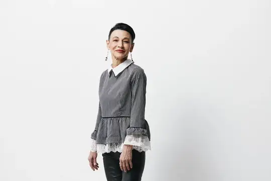 Модная азиатская женщина в черных кожаных брюках и серой блузке стоит у белой стены