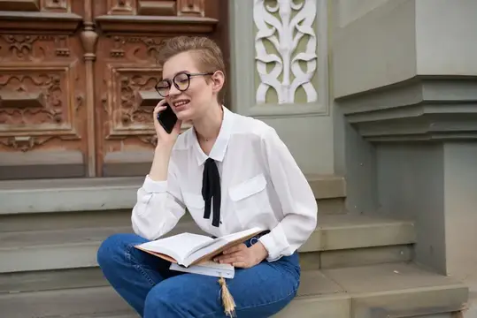 Улыбающаяся девушка в очках с книгой разговаривает по телефону на ступеньках