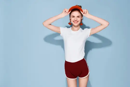 Улыбчивая девушка в белой футболке и красных шортах поправляет кепку