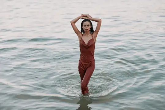 Молодая женщина в терракотовом комбинезоне стоит в воде с поднятыми руками
