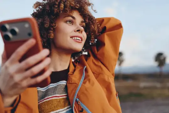 Кудрявая девушка в оранжевой куртке улыбается, глядя на смартфон