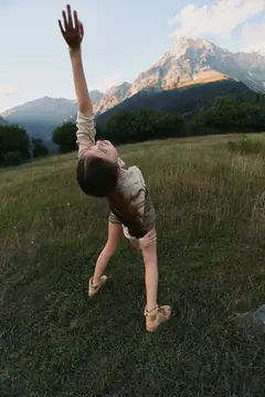 Яркая outdoor-сцена показывает женщину, наслаждающуюся природой: радостное движение по залитому солнцем полю с далекими пиками