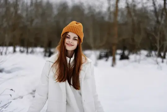 Рыжеволосая девушка в оранжевой шапке улыбается в зимнем лесу