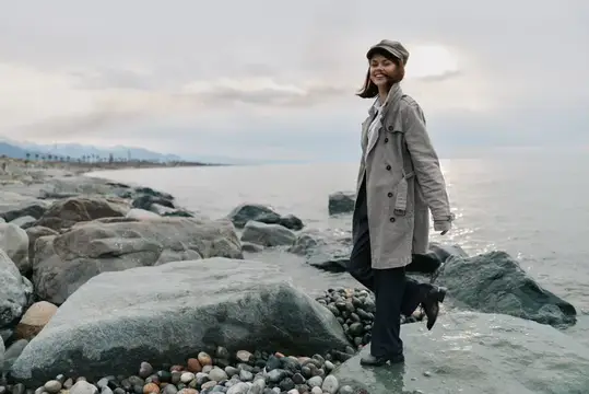 Улыбающаяся девушка в плаще и кепке стоит на камнях у моря