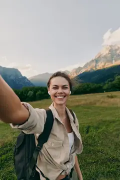 Улыбающаяся девушка с рюкзаком делает селфи на фоне гор