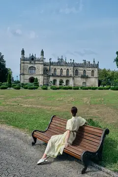 Девушка в светлом платье отдыхает на скамейке у старинного замка