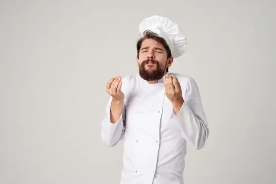Бородатый повар в колпаке жестом показывает вкусно на сером фоне