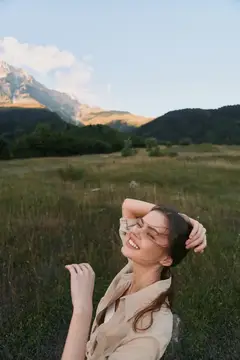 Жизнерадостная женщина стоит на открытом лугу на фоне гор, одетая в бежевую повседневную рубашку, наслаждаясь солнцем и ветерком