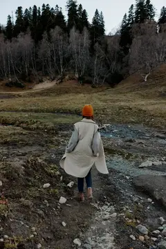Женщина в оранжевой шапке стоит спиной у горного ручья