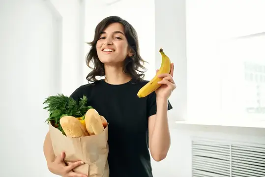 Улыбающаяся девушка с пакетом продуктов и бананом в руке